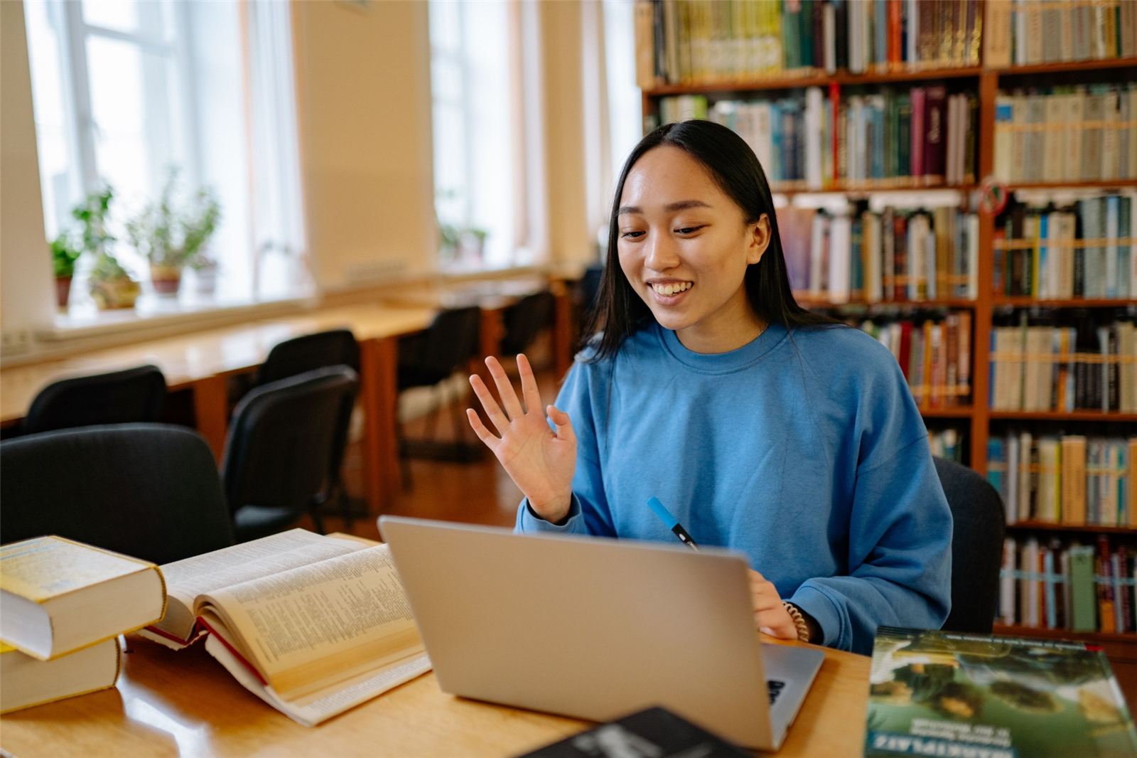 A smiling student waves during a video call with an advisor at a library table